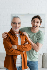 amazed young man hugging mature father in eyeglasses standing with crossed arms at home