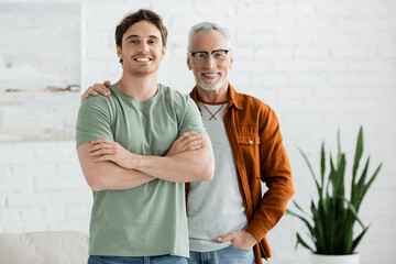 happy mature father in eyeglasses hugging cheerful young son posing with crossed arms at home