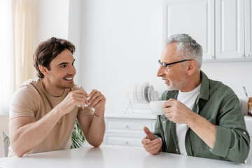 Obraz na płótnie Canvas happy young man drinking morning coffee with smiling and mature father at home