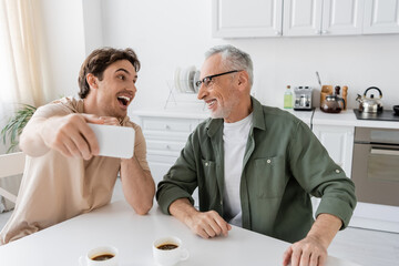 young excited man with mobile phone taking selfie with smiling father near coffee cups in kitchen - Powered by Adobe