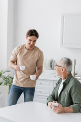 Fototapeta premium young man smiling while holding coffee cups near dad sitting with smartphone in kitchen