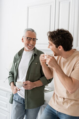 Fototapeta premium young man holding coffee cup and looking at cheerful dad during conversation in kitchen
