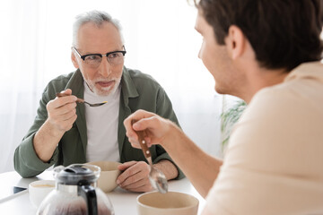 mature man in eyeglasses holding spoon and looking at blurred son during breakfast
