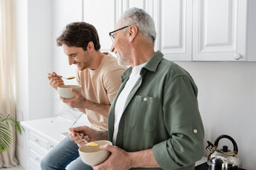 grey haired man looking at laughing son sitting on kitchen worktop with spoon and bowl of corn flakes