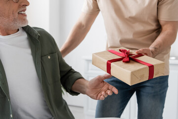 cropped view of young man holding gift box with red ribbon near smiling dad - obrazy, fototapety, plakaty