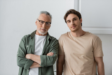 mature man in eyeglasses standing with crossed arms near young son and looking at camera