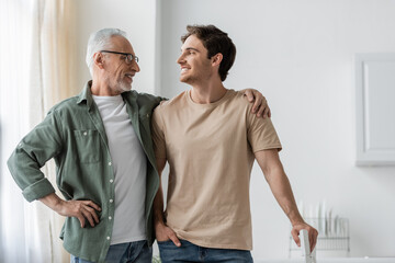cheerful grey haired man and young son looking at each other while posing in kitchen