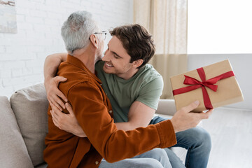 smiling man embracing dad holding fathers day present on couch in living room