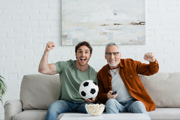 young excited man with mature dad showing win gesture near soccer ball and popcorn while watching championship on tv