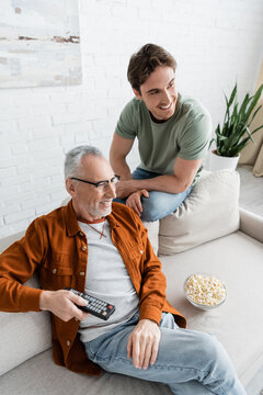 High Angle View Of Senior Man With Young Son Watching Tv On Couch Near Bowl Of Popcorn