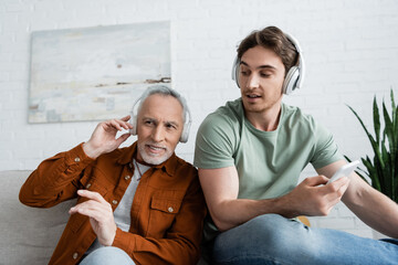 young man with mobile phone looking at mature dad smiling while listening music in headphones