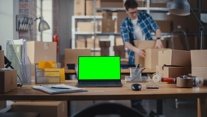 Desktop Computer Monitor Standing on a Table with a Green Screen Chromakey Mock Up Display. Small Business Storage Room with Worker Walking in the Background. Desk with Cardboard Boxes.