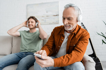 Fototapeta premium mature and happy man in wireless headphones using smartphone near son listening music on blurred background