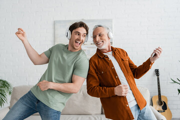 excited guy in wireless headphones singing and dancing with mature and cheerful dad at home