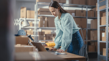 Small Business Owner of an Online Store Works on Laptop Computer while Standing at Her Desk in Warehouse. Female Employee Packing a Stylish Yellow Jumper in the Room with Shelves Full of Parcels.