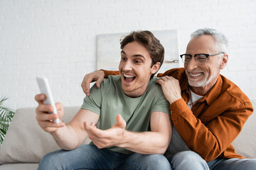 young man gesturing during video call while sitting with happy mature father in eyeglasses