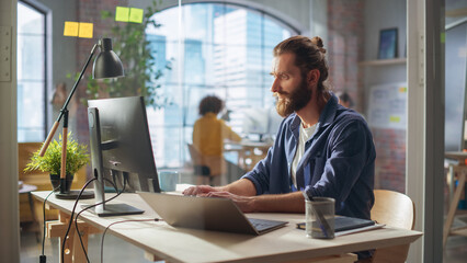 Stylish Long-Haired Bearded Specialist Sitting at a Desk in Creative Agency. Young Stylish Man Working on a Laptop Computer Connected to a Second Display. Office Team Members in the Background.