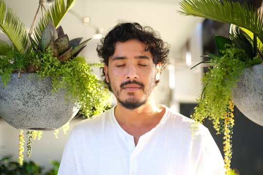 Curly haired man closing his eyes with hanging  plants behind him