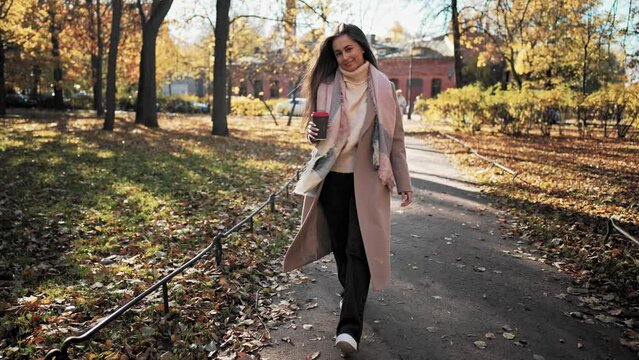 Full length front view young woman walking to camera with confident step and gait, in autumn park with mug of coffee in hands. Outdoors nature landscape. Warm drink and cozy clothes in cold weather.