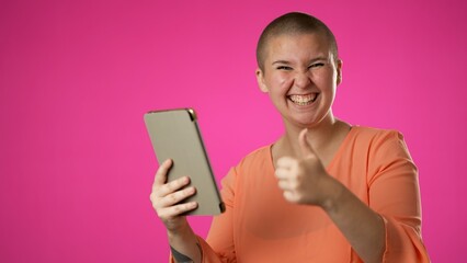 Portrait of happy smiling gender fluid non binary woman promoter 20s shopping online on mobile tablet computer giving thumbs up isolated on pink background studio with copy space