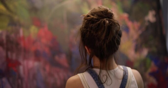 Back of head of artist woman with brown hair in bun who paints abstract picture on huge canvas. Professional female artist turns around and looks at camera against background of an abstract painting