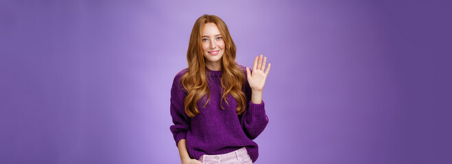 Shy and cute redhead female newbie greeting new team smiling friendly and waving raised palm in hello or hi gesture, saying nice to meet you as getting know new member over violet wall - obrazy, fototapety, plakaty