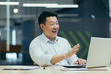 Business online meeting. A young asian businessman holds a meeting with partners, clients, a conference. Sitting at the desk in the office, looking at the laptop, explaining, smiling.