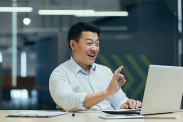 A young Asian businessman conducts an online interview for a job in company. He sits in the office at a table with a laptop, talks through a video call, points with his fingers ok, smiles.