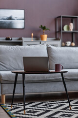 Vertical background image of open laptop by comfortable couch in elegant home interior with mauve wall