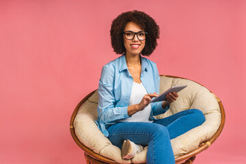 Portrait of young african american woman with curly african hair holding digital tablet and smiling standing over isolated pink background with copy space.