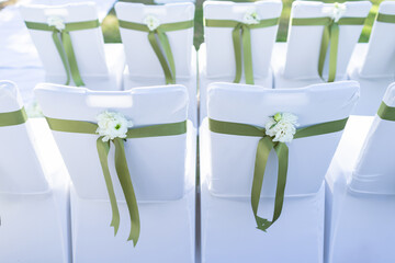 White chairs set up in a row at the ceremony There was an ornamental flower attached to the back, and a green cloth was tied around it.