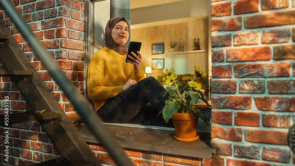 Aufkleber portrait of a young arab woman using a smartphone while sitting on a window sill in a multi-storey b