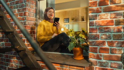 Portrait of a Young Arab Woman Using a Smartphone while Sitting on a Window Sill in a Multi-Storey...