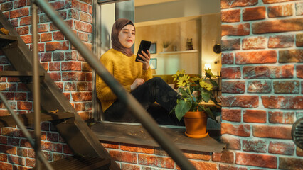 Muslim Girl Wearing a Hijab, Using a Smartphone while Sitting on a Window Sill in Brooklyn Style...