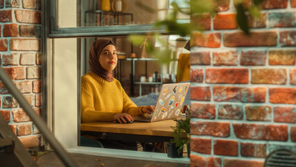 Young Muslim Female Student Using Laptop at Home. Attending Remote Education During the Day. Working on Computer, Writing an Essay Assignment. Arc Shot from Outside the Window.