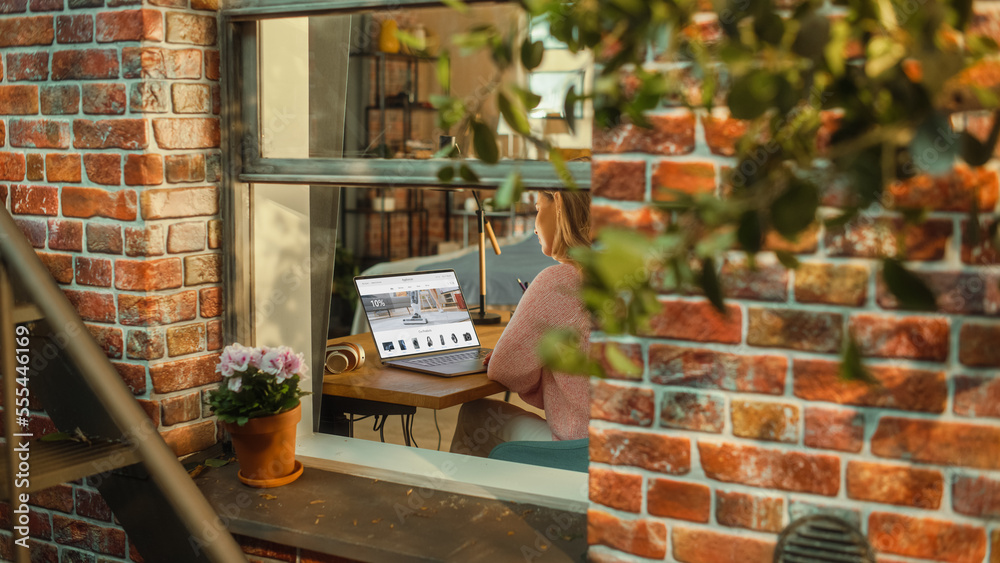 Poster Female Working on a Laptop Computer with Online Shopping Website Display while Sitting Behind a Table in her Living Room. Business Manager Working Remotely from Home. View from Outside the Window.