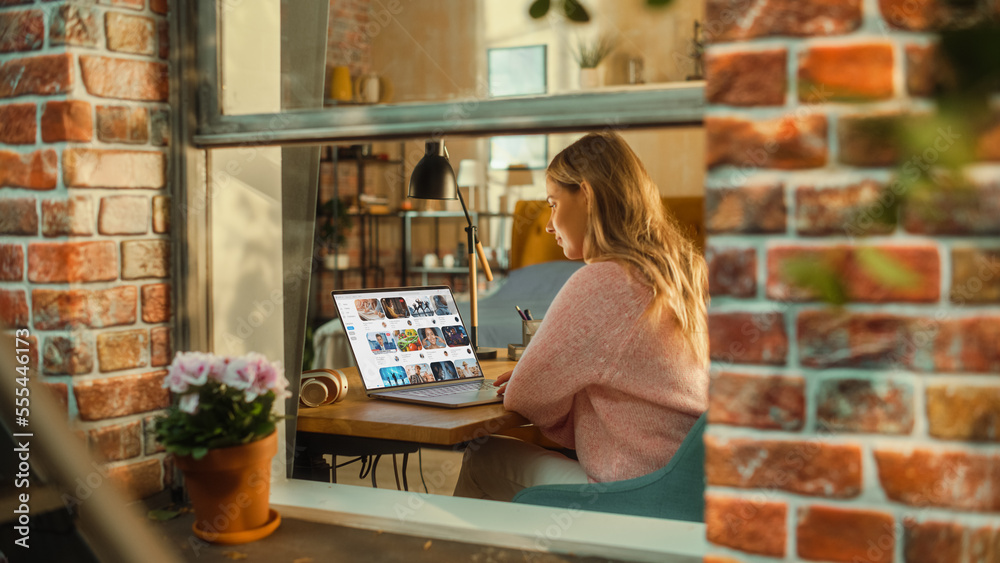 Poster Female Working on a Laptop Computer with Video Platform Display while Sitting Behind a Table in Stylish Living Room. Marketing Manager Working Remotely from Home. View from Outside the Window.