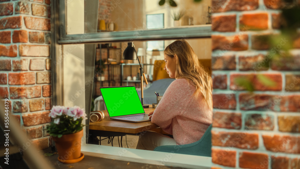 Aufkleber Female Working on a Laptop Computer with Green Screen Mock Up Display while Sitting Behind a Table in Stylish Living Room. Business Manager Working Remotely from Home. View from Outside the Window.