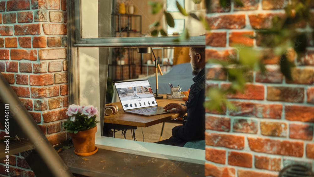 Aufkleber Black Man Using Laptop Computer with Online Shopping Website Display while Sitting Behind a Table in Stylish Living Room. Husband Shopping for Appliances Using Internet. Shot from Outside the Window.