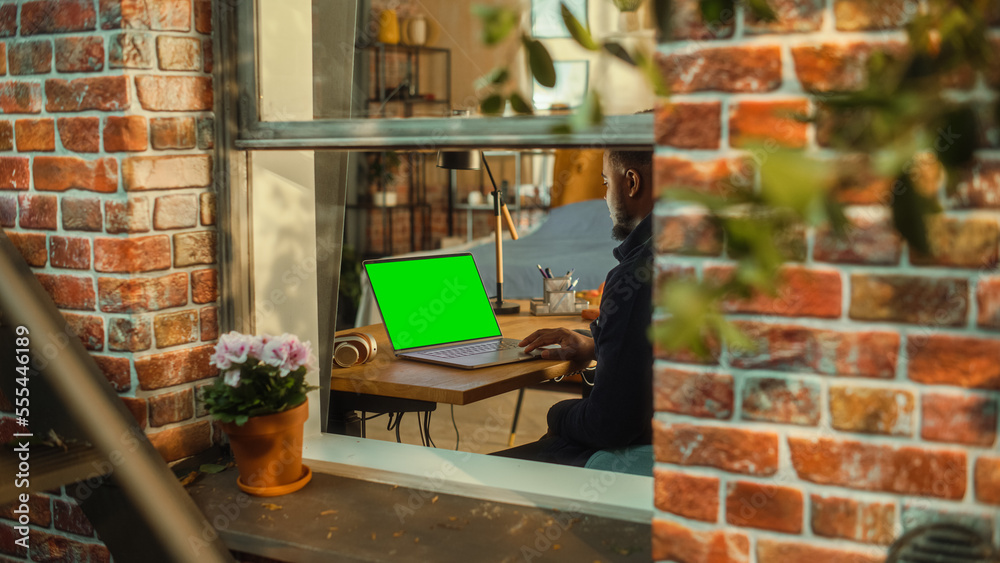 Poster African Man Working on a Laptop Computer with Green Screen Mock Up Display while Sitting at a Table in Stylish Living Room. Business Manager Working Remotely from Home. View from Outside the Window.