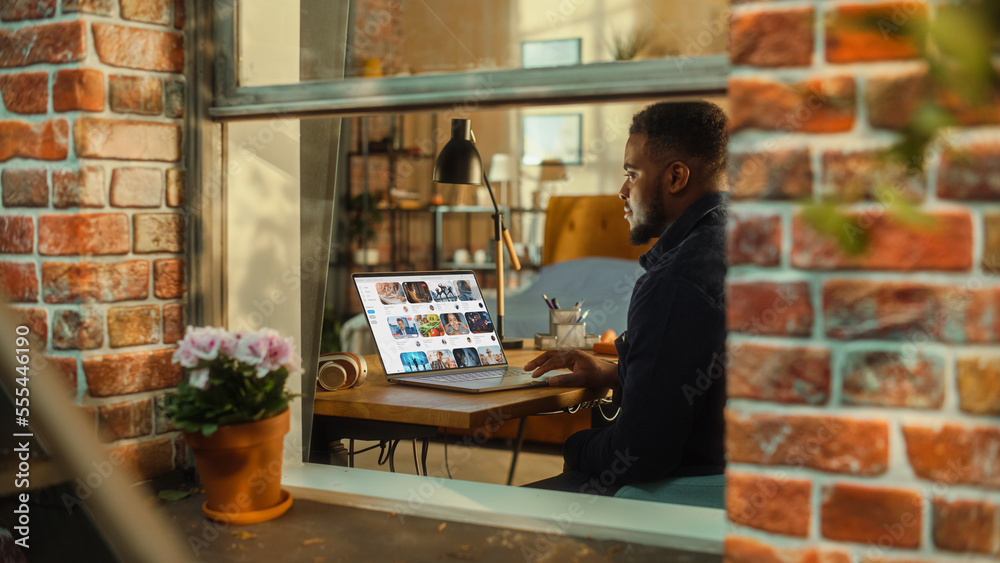 Poster Black Man Using Laptop Computer with Video Streaming Platform Display while Sitting Behind a Table in Stylish Living Room. Influencer Browsing Internet. Shot from Outside the Window.