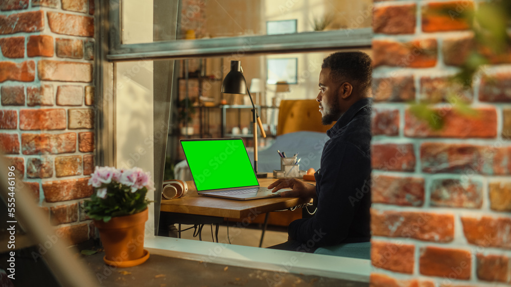 Aufkleber African Man Working on a Laptop Computer with Green Screen Mock Up Display while Sitting at a Table in Stylish Living Room. Business Manager Working Remotely from Home. View from Outside the Window.