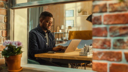 Young Handsome Male Using Laptop Computer, Sitting in Living Room in Apartment. African American...