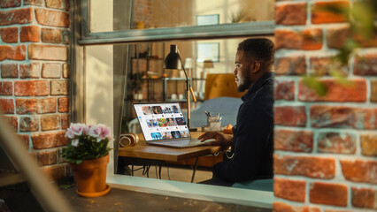 Black Man Using Laptop Computer with Video Streaming Platform Display while Sitting Behind a Table in Stylish Living Room. Influencer Browsing Internet. Shot from Outside the Window.