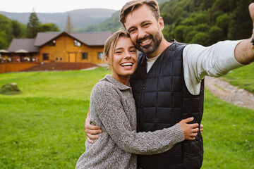 Young couple taking selfie and hugging while standing outdoors with house on the background