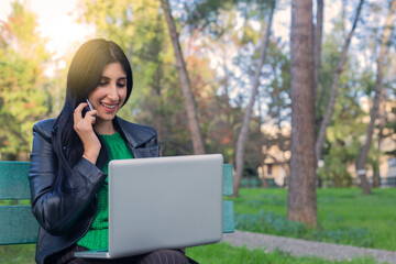 Single student on line with a laptop and calling customer service on the phone lying on a bench in the street with a green background