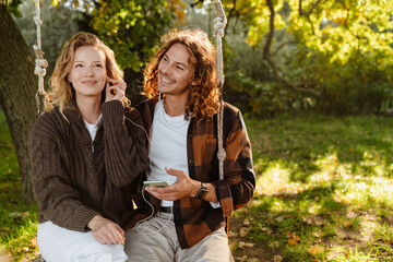 White happy couple listening to music while sitting on swing in park