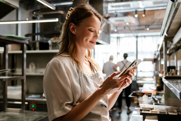 Smiling woman cook using mobile phone while standing in kitchen of a restaurant