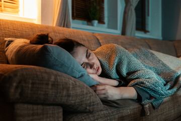 Flu and desease. Illness young Caucasian woman lies on sofa covered with blanket and watching tv....