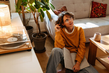 Young white woman in eyeglasses reading book while sitting on floor
